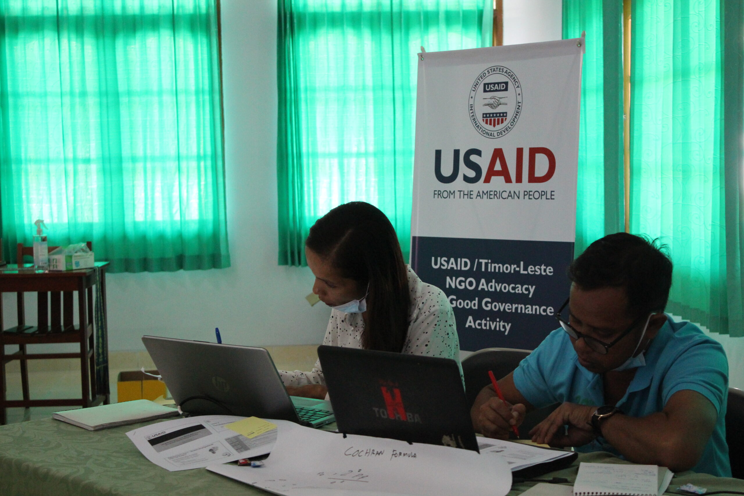 Moniz and a colleague working at computers at a training in front of USAID banner.